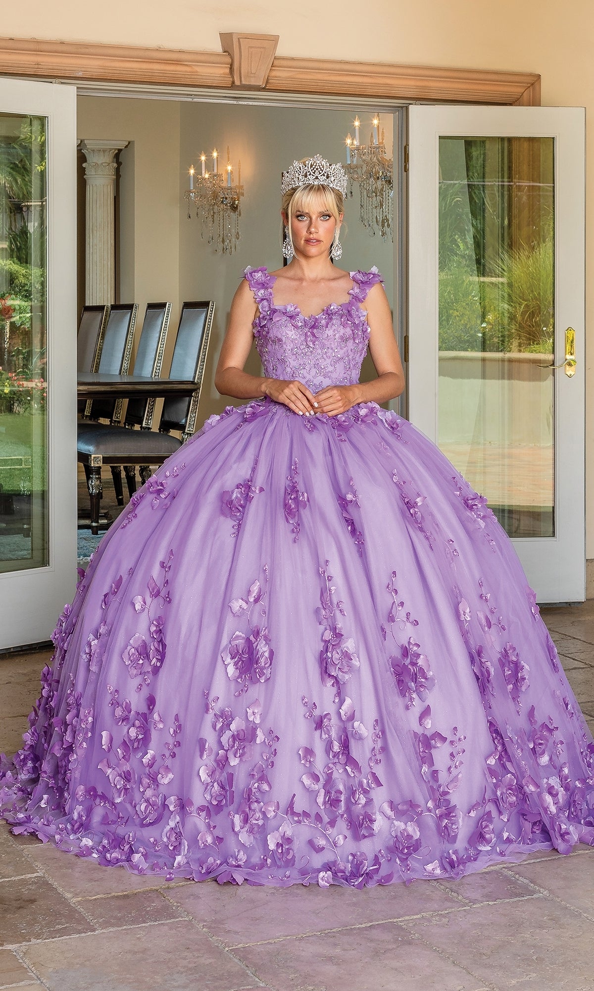 A person in the Quinceanera Dress 1613 By Dancing Queen, featuring lavender floral appliqués and off-the-shoulder details, stands by open glass doors with a chandelier and dining table in the background.